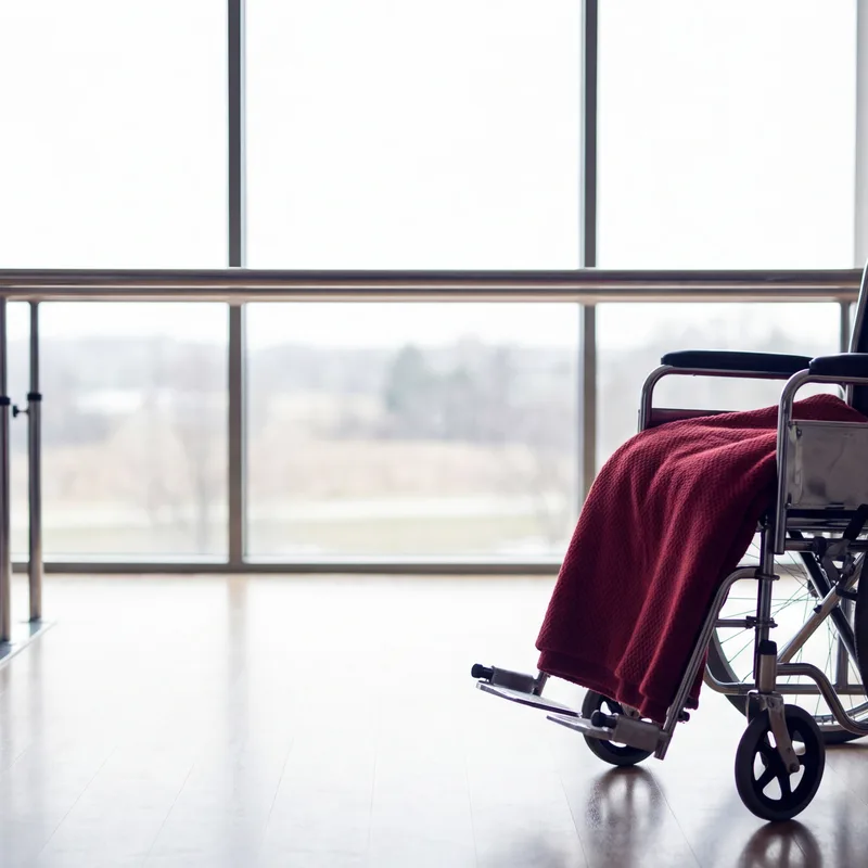 Rehabilitation room with parallel bars leading to a bright window and a wheelchair with a crimson blanket
