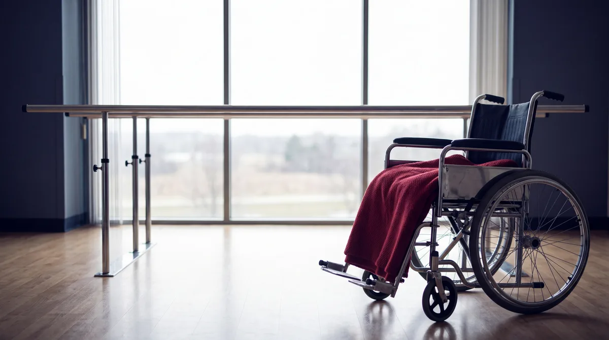 Rehabilitation room with parallel bars leading to a bright window and a wheelchair with a crimson blanket