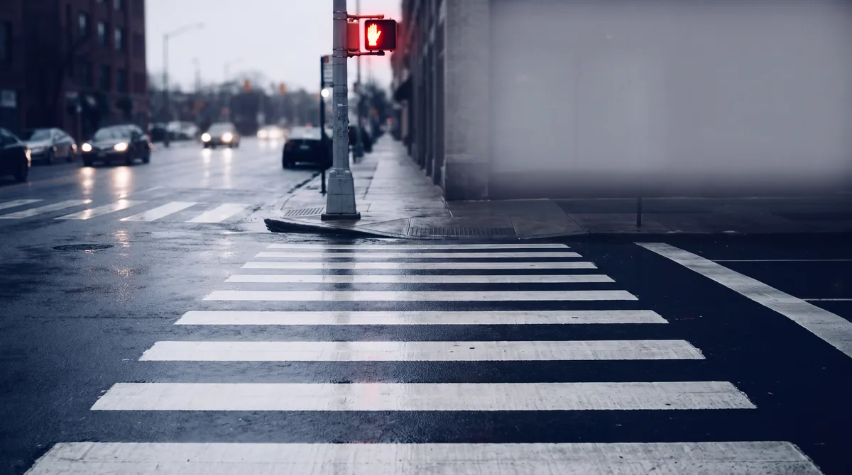 Crosswalk at dusk with approaching headlights and a crimson pedestrian signal